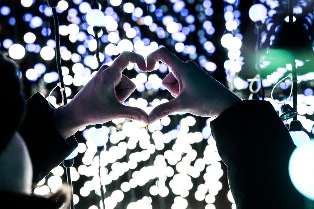 A hand forms a heart with fairy lights in the background.